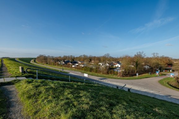 Groen weiland met een dijk en een kronkelige weg langs huizen in de achtergrond.