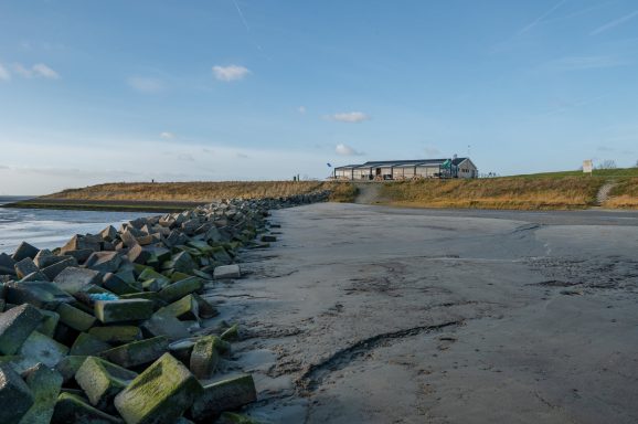 Uitzicht op een strand met een kade en een gebouw in de achtergrond onder een blauwe lucht.