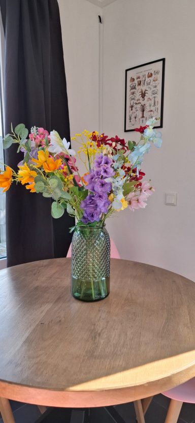 Een vaas met kleurrijke bloemen staat op een houten tafel in een lichte ruimte.